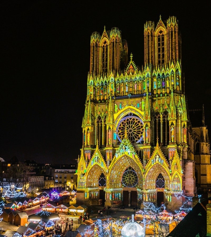 Façade occidentale de la cathédrale Notre-Dame de Reims, chef-d’œuvre gothique des sacres des rois de France inscrit au patrimoine mondial de l’UNESCO, illuminée de projections colorées à la nuit tombée, avec sa grande rose, ses trois portails sculptés et ses deux tours, au-dessus des chalets illuminés du marché de Noël sur le parvis, dans le département de la Marne, en région Grand Est. Façade occidentale de la cathédrale Notre-Dame de Reims, chef-d’œuvre gothique des sacres des rois de France inscrit au patrimoine mondial de l’UNESCO, illuminée de projections colorées à la nuit tombée, avec sa grande rose, ses trois portails sculptés et ses deux tours, au-dessus des chalets illuminés du marché de Noël sur le parvis, dans le département de la Marne, en région Grand Est.