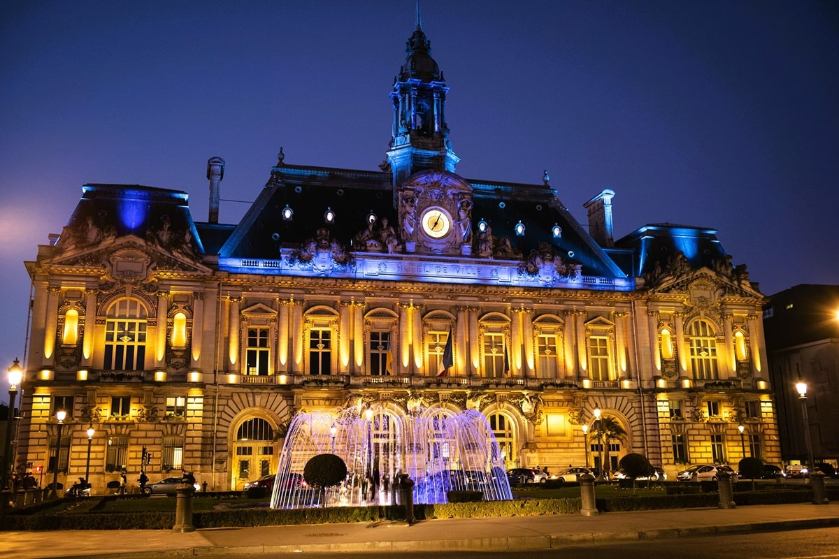 Hôtel de ville de Tours illuminé à la nuit tombée, œuvre de l’architecte Victor Laloux et monument historique, avec son campanile, son horloge encadrée des cariatides du Jour et de la Nuit et les jets d’eau de la fontaine de la place Jean-Jaurès, dans le département d’Indre-et-Loire, en région Centre-Val de Loire.